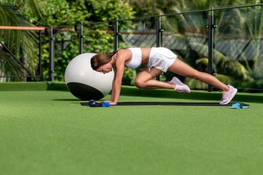 Güzel, ince bir kız güneş gözlüğü takıyor, spor üniforması giyiyor karın kasları için egzersiz yapıyor, barda bacağını karnına çekiyor. Spor egzersizleri..