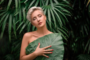 Kapalı gözlü güzel sarışın manken. Doğal makyaj ve yeşil yapraklar. Makyajlı, kırbaçlı, kaplıca ve sağlıklı genç kadın cilt bakımı konsepti. Kapat.,