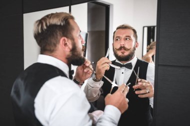 Ciddi yakışıklı, şık bıyıklı ve sakallı bir adam elinde bir makas ve elinde bir ayna tutuyor ve aynaya hayran.