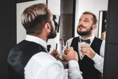 Şık bıyıklı ve sakallı genç bir adam elinde bir makas ve aynayla aynada hayranlık duyuyor.