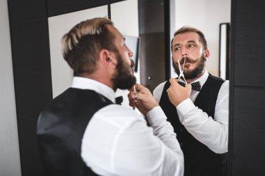 Lüks bıyıklı ve sakallı yakışıklı adam onları aynanın önünde makasla kesiyor.