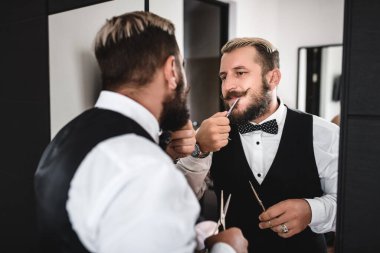 Tarz sahibi bir adam aynanın önünde çelik bir bıçakla sakalını kesiyor.