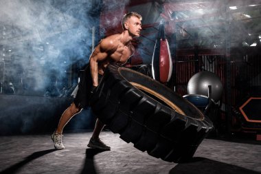 Genç kaslı bir adam spor salonunda ağır lastik çeviriyor.