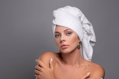 Kafasına beyaz havlu geçirmiş, gri arka planda çıplak duran beyaz bir kadının stüdyo fotoğrafı. Cilt bakımı, kozmetik ürün konsepti