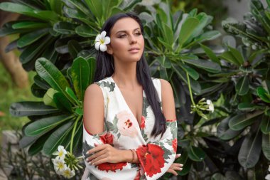 Saçında beyaz çiçekle yeşil yaprakların arkasında poz veren çekici bir kadın.