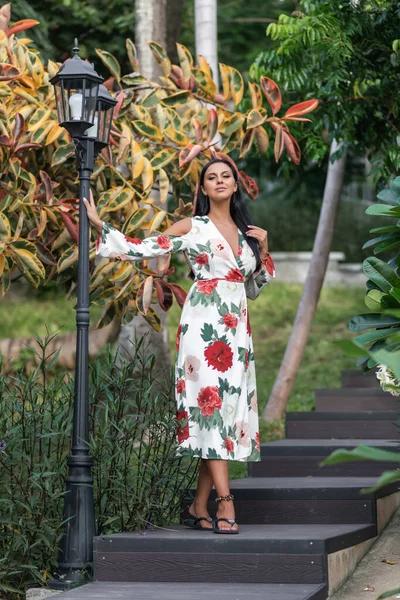 Beautiful woman with black hair wearing summer white dress with flowers print, posing in the street. Woman stands near the lantern.