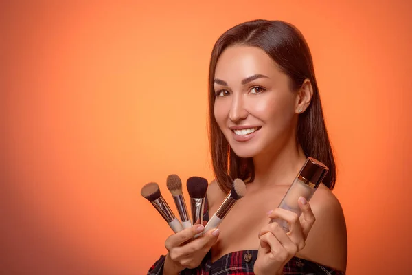 Beautiful young woman with makeup brushes and foundation in her hands. Professional makeup artist. Studio background