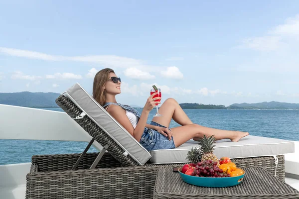 Side view: a charming woman lies on a sunbed, drinking watermelon juice while sailing on a yacht during a summer sea cruise. Tropical summer fruits on a plate next to her.