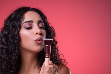 Pudra fırçasında esen çekici genç bir kadın. Parlak kozmetik ürünleri. Profesyonel makyaj malzemesi. Pembe stüdyo geçmişi. Portre.