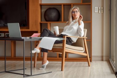 Lüks bir iş kadını elinde kahveyle masada oturan bir dizüstü bilgisayarda çalışıyor. Serbest, uzak çalışma.