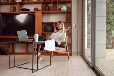 Beyaz gömlekli bir kadın çalışma masasında çalışırken elinde bir telefonla oturuyor.