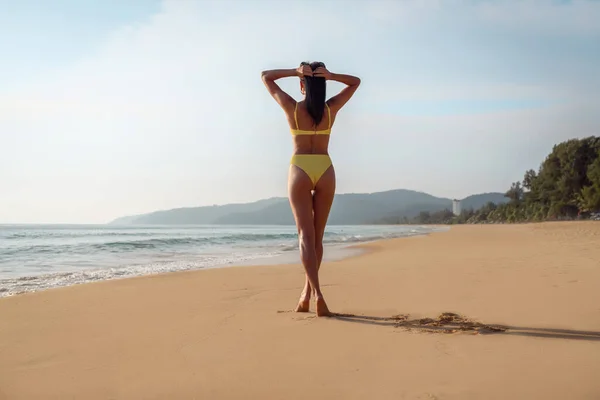 View from behind: A slender girl in a yellow bikini posing near the sea