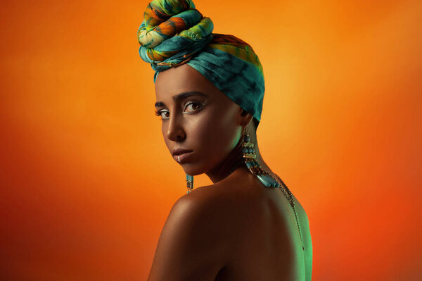 Beauty bright oriental woman with creative make up, shawl on head. Orange studio background