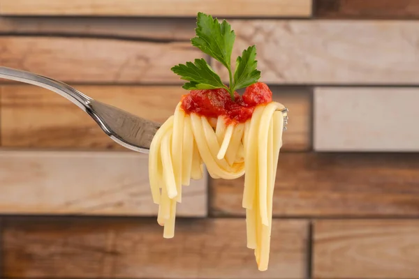 spaghetti on a fork with tomato sauce and parsley leaf close-up on a blurred wooden background