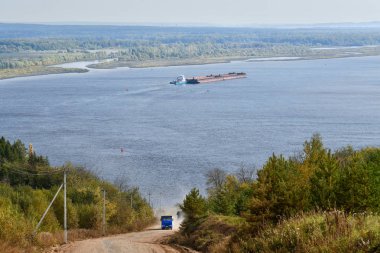 Bir damperli kamyon toprak bir yol boyunca yokuş yukarı yükselir. Arka planda büyük bir nehir var..