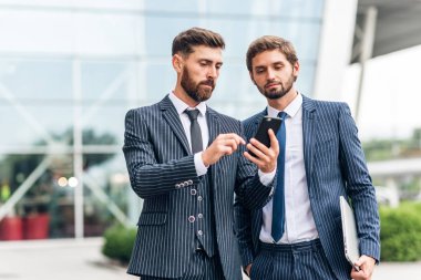 Takım elbiseli genç işadamı cep telefonu kullanarak ve onlar modern binanın yanında açık havada ayakta iken meslektaşına bir şey gösteren