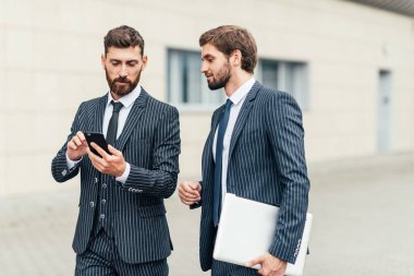 İki çekici gülümseyen genç ofis arkadaşları şehir sokaklarında açık havada duran takım elbise giyiyor, cep telefonu bakarak