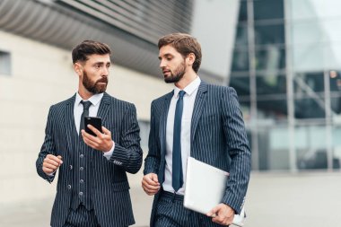 İki çekici gülümseyen genç ofis arkadaşları şehir sokaklarında açık havada duran takım elbise giyiyor, cep telefonu bakarak