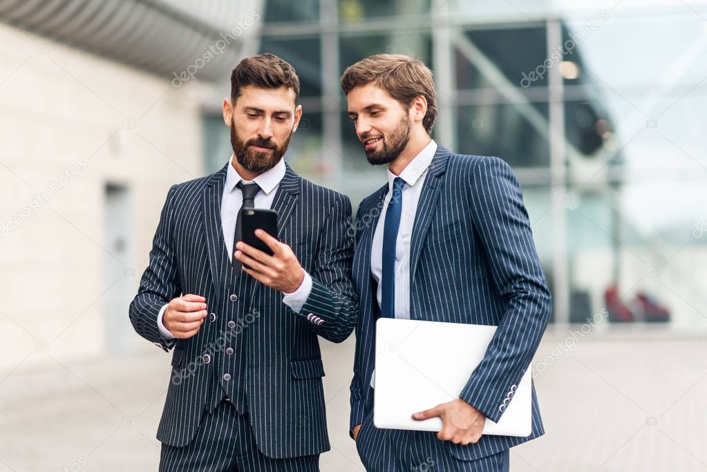 Two attractive smiling young businessmen wearing suits walking outdoors at the city streets, , using mobile phone
