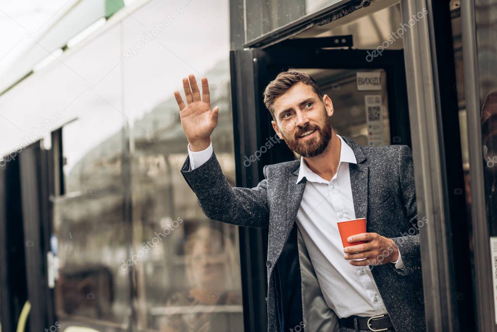Positive man waving hand while calling colleague outdoors. Jolly handsome young businessman in greeting friend and standing on stairs. Meeting in city concept