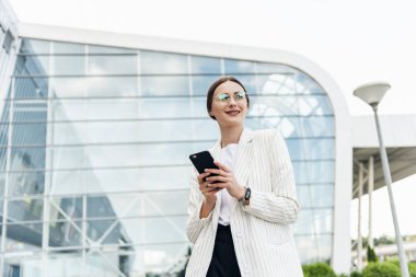 Ofis yakınında cep telefonu kullanarak Mutlu güzel esmer kız, güzel kadın telefon tarama ve alışveriş merkezi yakınında yürürken gülümseyerek bir mobil uygulama online alışveriş yapıyor.