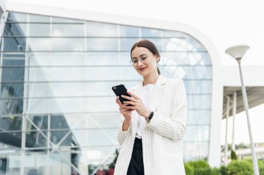Ofis yakınında cep telefonu kullanarak Mutlu güzel esmer kız, güzel kadın telefon tarama ve alışveriş merkezi yakınında yürürken gülümseyerek bir mobil uygulama online alışveriş yapıyor.