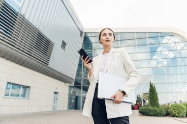 İşini bir yolla yapabilir. Açık havada dururken gülümseyen dizüstü bilgisayarını kullanan tam uzunlukta ki genç iş kadını.