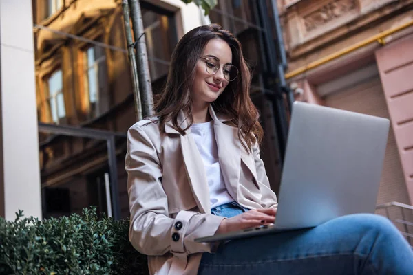 Close-up of young professional businesswoman using her laptop outside while working at modern business center, modern buildings in the background