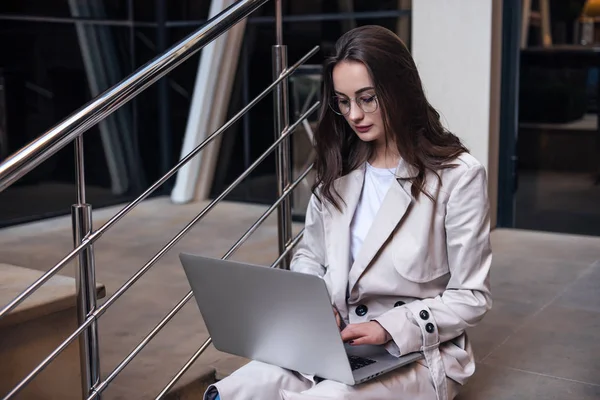 Close-up of young professional businesswoman using her laptop outside while working at modern business center, modern buildings in the background