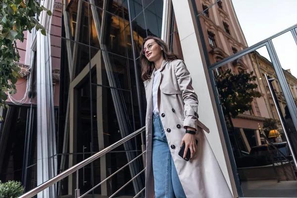 Güneş gözlüklü, gülümseyen kıvırcık kadın Central City caddesinde yürüyor ve telefonunu kullanıyor. Beyaz ceketli güzel yaz kadını caddede yürürken cep telefonuna bakıyor.