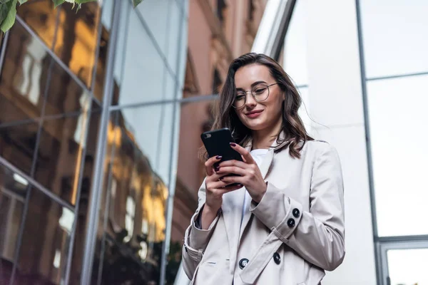 Smiling curly woman wearing trendy sunglasses walks down the central city street and uses her phone. Pretty summer woman in white jacket walks down the street looking at her mobile phone