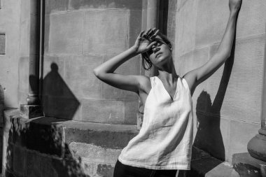 Beyaz tişörtlü bir kadın sokakta dikiliyor, yüzünü gölgeli güneşten koruyor..