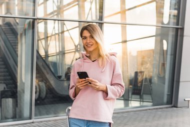 Havalimanında dikilirken akıllı telefon kullanan gündelik giysili genç sarışın kadın turist..