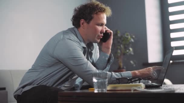 Бізнесмен працює на сенсорній панелі комп'ютера, розмовляючи на смартфоні, молодий бізнесмен використовує ноутбук, сидячи за дерев'яним столом . — стокове відео