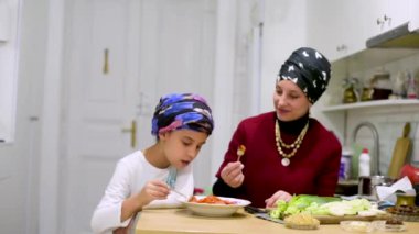 Anne ve kızı yemek masasında taze sebze salatası yiyor. Anne ve kızı mutfakta yemek yiyorlar. Karantina karantinasında sosyal uzaklık ve kişisel izolasyon.
