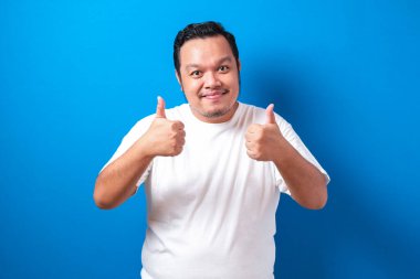 Beyaz tişörtlü şişman Asyalı adam başparmağını kaldırırken kameraya gülümsüyor. Mavi arkaplana karşı seçici odaklı yarı vücut portresi.