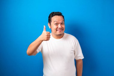 Beyaz tişörtlü şişman Asyalı adam başparmağını kaldırırken kameraya gülümsüyor. Mavi arkaplana karşı seçici odaklı yarı vücut portresi.