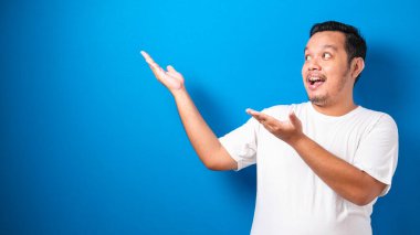 Beyaz tişörtlü komik Asyalı bir adamın portresi gülümsüyor ve mavi arkaplanda fotokopi alanı olan bir şeyi gösteriyordu.