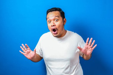 Beyaz tişörtlü şişman Asyalı adam mavi arka planda komik ve sürpriz bir ifade sergiliyor.