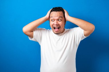 Beyaz tişörtlü şişman Asyalı adam mavi arka planda komik ve sürpriz bir ifade sergiliyor.