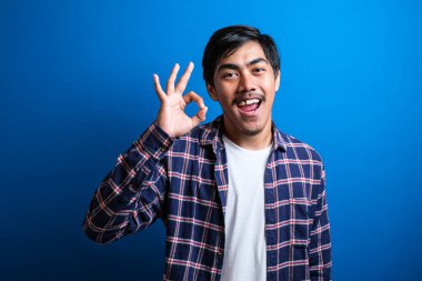 Asyalı genç bir üniversite öğrencisi tamam tabelasıyla gülümsüyor. Mavi arka planda mutlu Asyalı öğrenci..