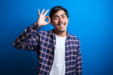 Asyalı genç bir üniversite öğrencisi tamam tabelasıyla gülümsüyor. Mavi arka planda mutlu Asyalı öğrenci..