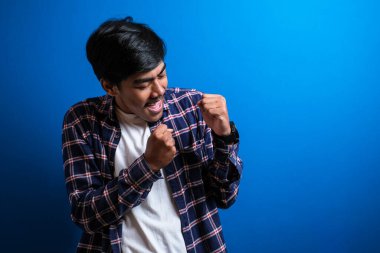 Mavi arka plan stüdyosunda izole edilmiş neşeli, genç bir öğrenci zaferini kutlamak için kolunu kaldırdı. Adam aldığı iyi haberle mutlu bir şekilde dans ediyor..