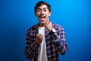 Mavi arka plan stüdyosunda izole edilmiş neşeli, genç bir öğrenci zaferini kutlamak için kolunu kaldırdı. Adam aldığı iyi haberle mutlu bir şekilde dans ediyor..