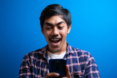 Genç Asyalı öğrenci gülümsüyor ve mavi arka planda izole edilmiş telefonu kullanıyor. Adam arkadaşıyla sohbet etmekten mutlu görünüyor.