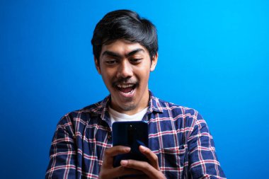 Genç Asyalı öğrenci gülümsüyor ve mavi arka planda izole edilmiş telefonu kullanıyor. Adam arkadaşıyla sohbet etmekten mutlu görünüyor.