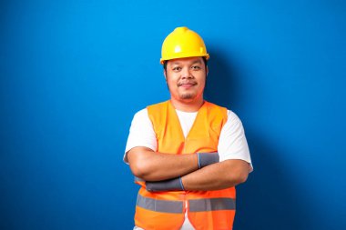 Kasklı ve güvenlik yeleği giyen şişman Asyalı işçi potansiyeli mavi arka plana karşı güven jesti yaparak elini çaprazladı.