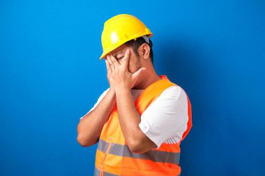 Üniformalı ve kasklı şişman Asyalı inşaat işçisi izole edilmiş mavi arka planda gözleri ellerle kaplıyor ve üzgün ve korku ifadeleriyle dur hareketi yapıyor. Utanç verici ve olumsuz bir kavram..