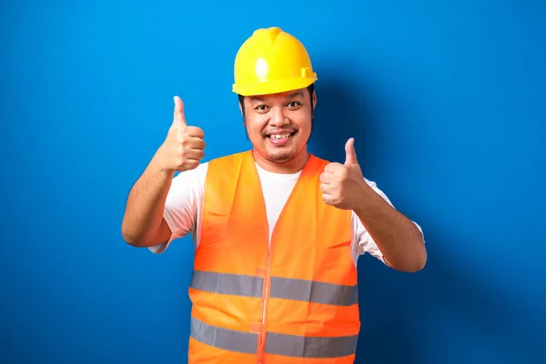 Fat asian construction worker wearing orange safety vest and helmet showing thumb up sign smiling on blue background