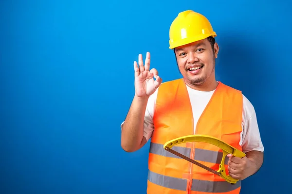 Fat asian construction worker wearing orange safety vest and helmet showing okay sign while holding hacksaw on blue background